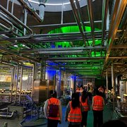 Teilnehmende in orangefarbenen Warnwesten gehen durch einen Produktionsbereich der Brauerei Weihenstephan mit Edelstahlbehältern und zahlreichen Rohrleitungen.